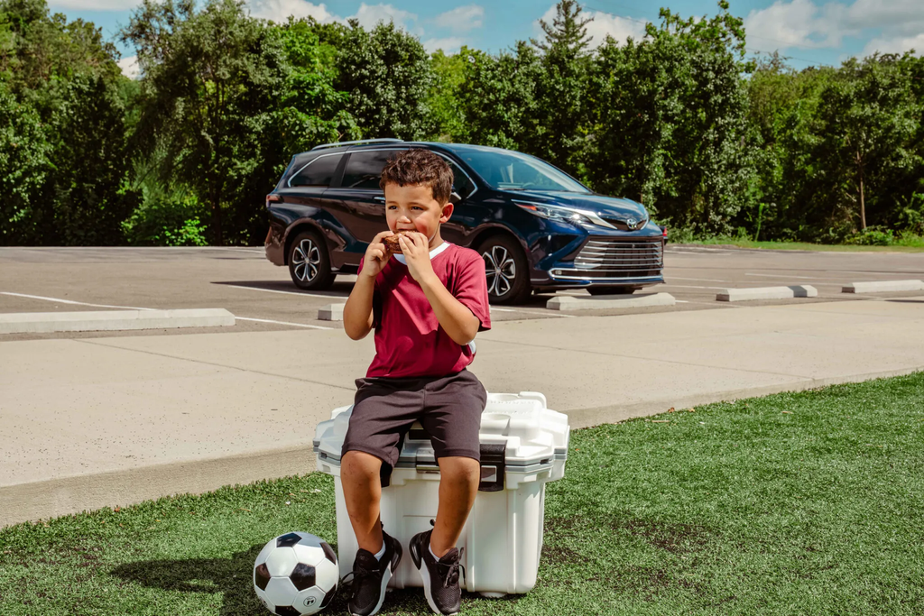 Boy eating a Chip Wheels ice cream sandwich on a cooler beside a soccer ball with a Toyota Sienna parked behind